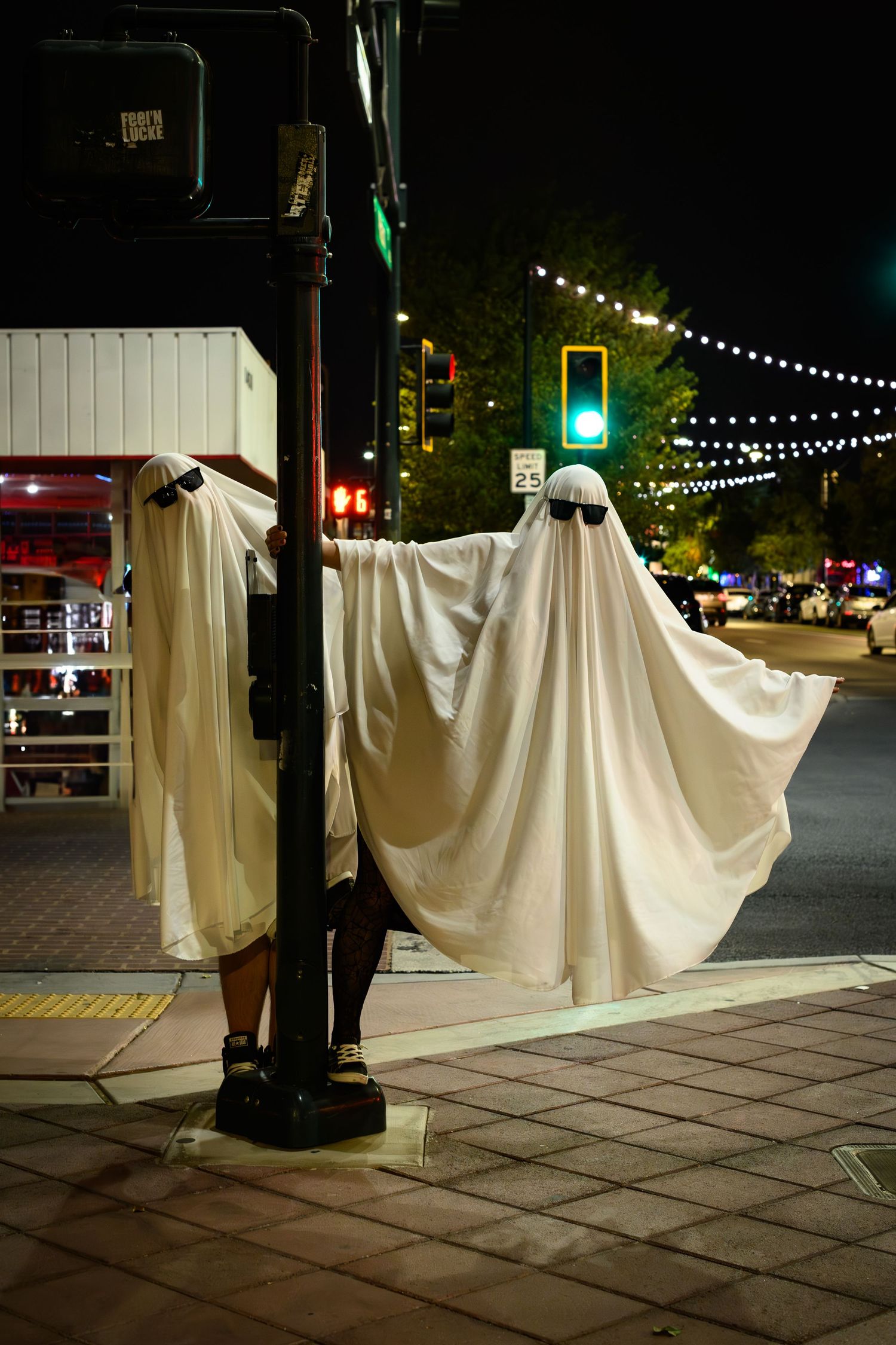 White ghost-like Halloween decorations hang between posts on a dark city sidewalk with string lights overhead.
