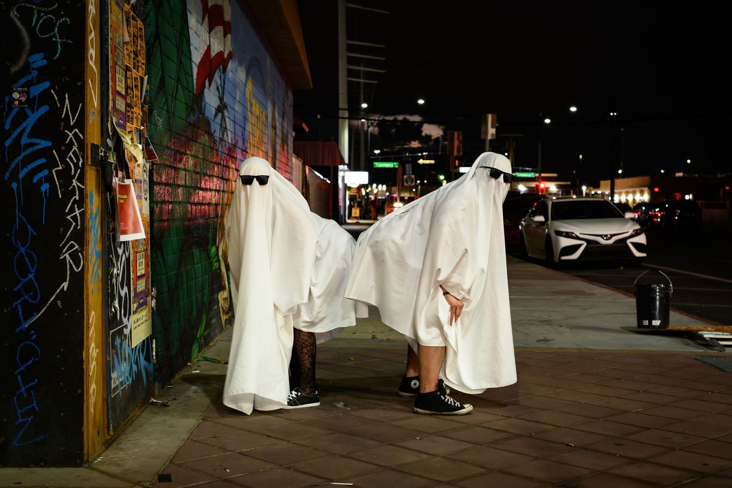 People dressed as ghosts in white sheets walk through dark city streets at night creating an eerie Halloween atmosphere.