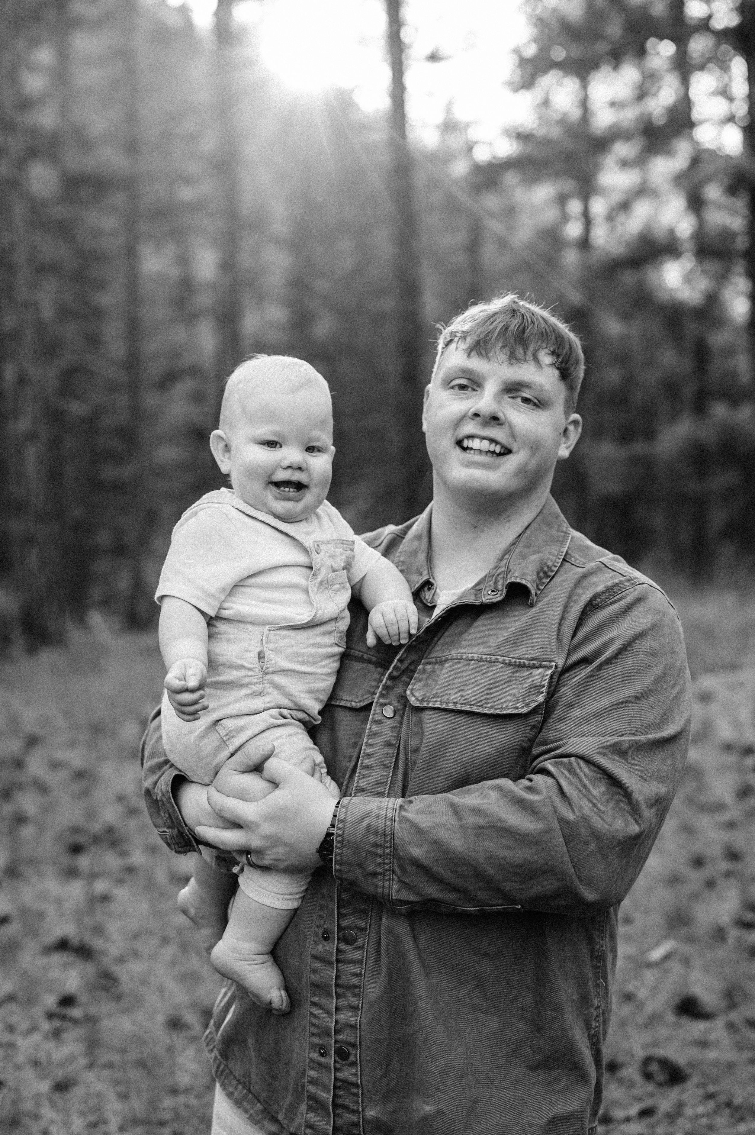 A heartwarming black and white portrait taken in a sunlit forest setting.