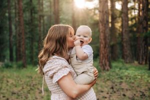 A tender moment captured in a sunlit forest as a parent kisses a joyful baby wearing light colored clothing.