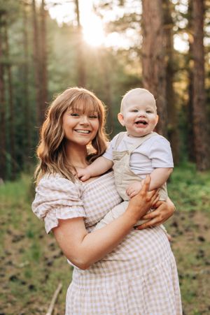 A heartwarming forest photoshoot showing a parent and baby sharing smiles in golden sunlight.