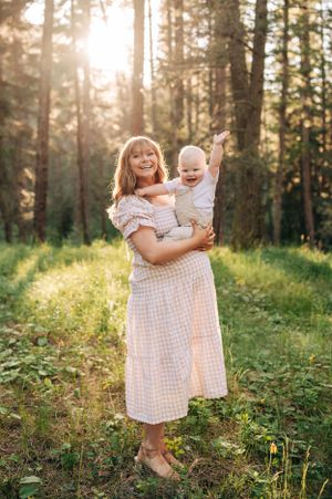 Playful moment in a pine forest as a parent lifts a laughing baby while wearing a checkered dress.