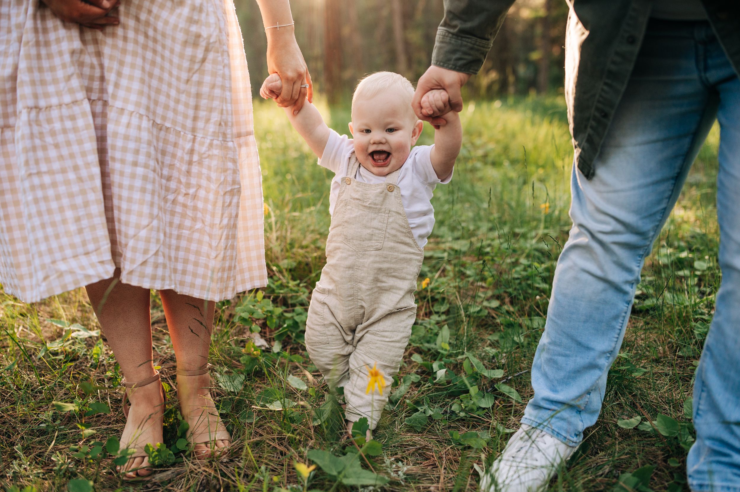 Baby taking first steps while holding hands with parents in a forest clearing.