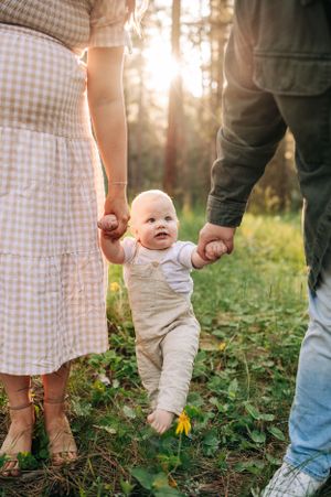 Baby learning to walk while holding both parents' hands in a sunlit forest setting.