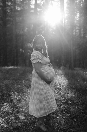 Silhouette of an expectant parent in a gingham dress standing in dramatic forest lighting.