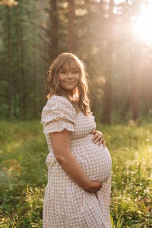 Maternity portrait in a forest with golden sunlight highlighting a gingham dress.