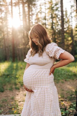 Side view of an expectant parent in a light gingham dress posing in dappled forest light.