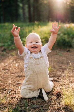 Joyful baby in beige overalls raises arms while sitting on forest ground at sunset.
