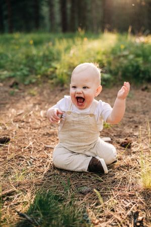 Excited baby in beige overalls sits on ground with arms raised in sunlit forest.