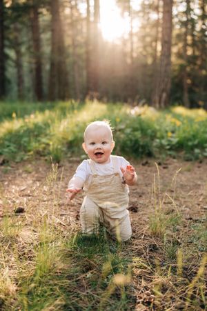 Happy baby in overalls taking first steps in a sunlit forest clearing.