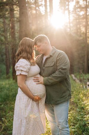 Tender moment between expectant couple in forest with golden sunlight streaming through trees.