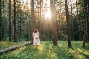 Expectant mother in gingham dress poses in sunlit forest clearing.