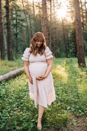 Woman in checkered dress cradles baby bump while standing in forest.