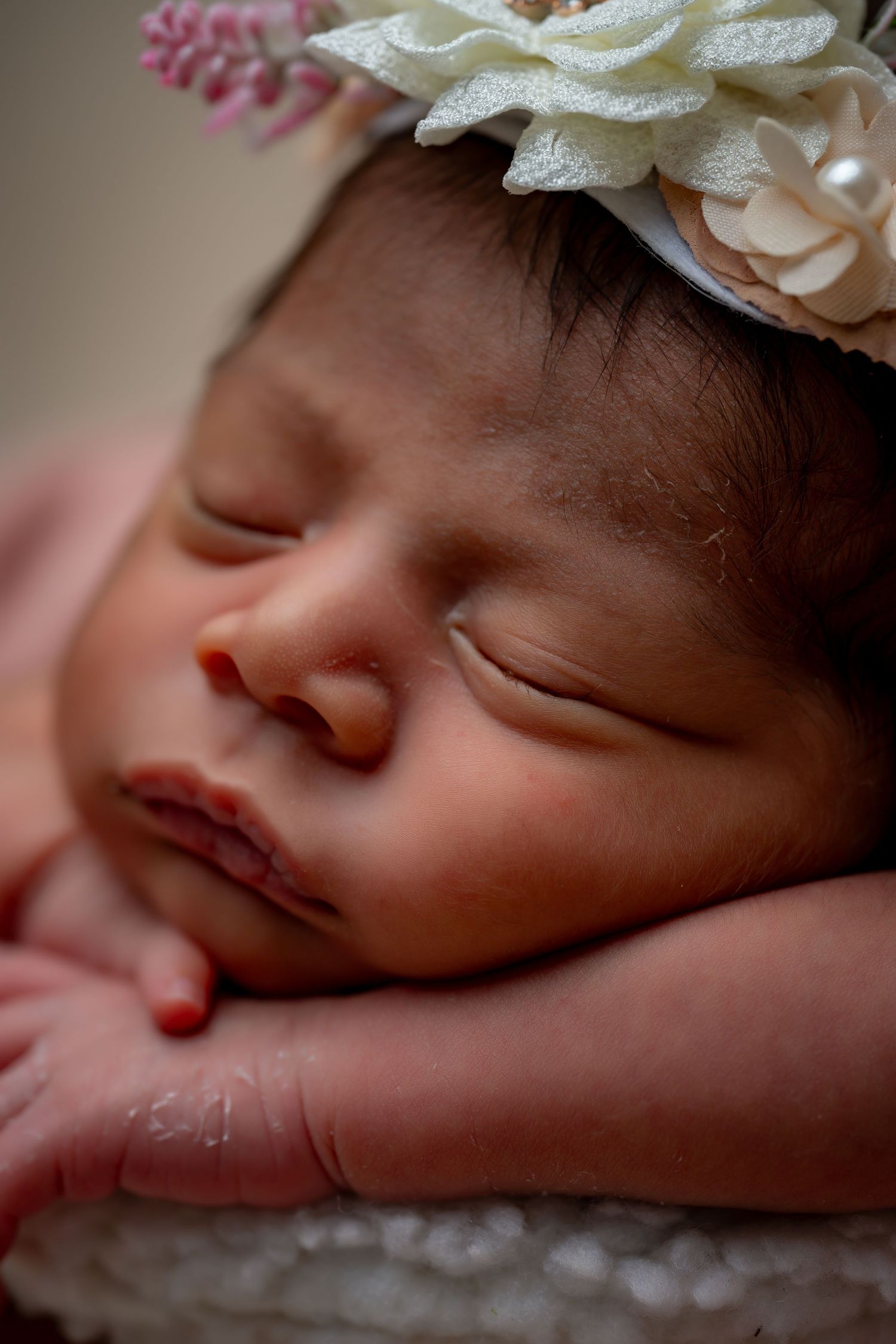 Close-up portrait of a sleeping newborn wearing a white floral headband against a soft pink background.