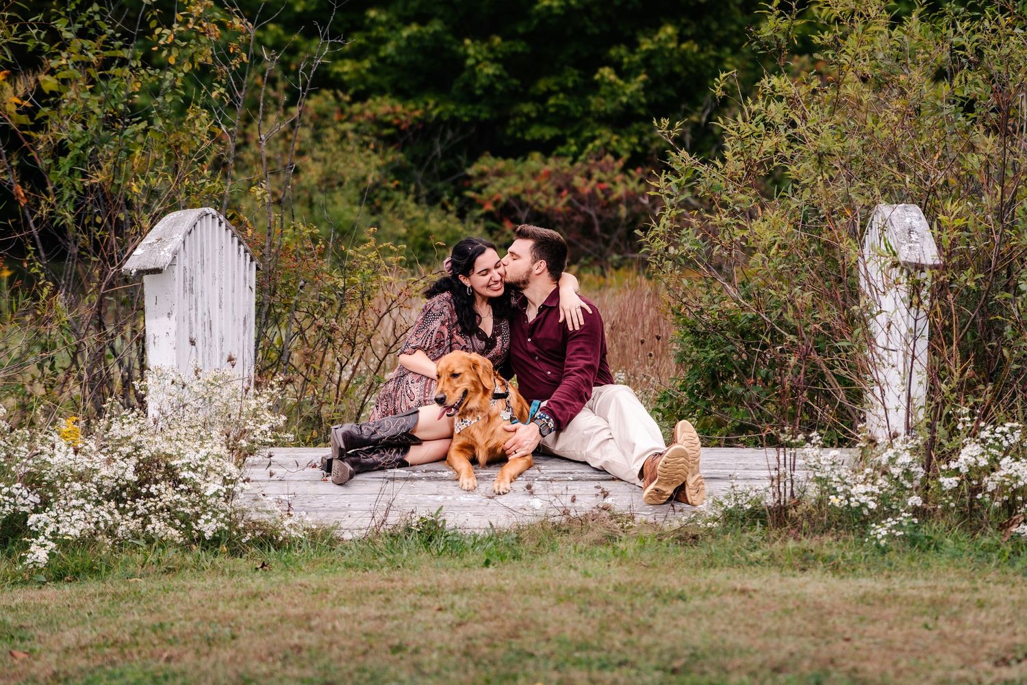 Couple having intimate picnic with their dog between white fence posts in autumn field.