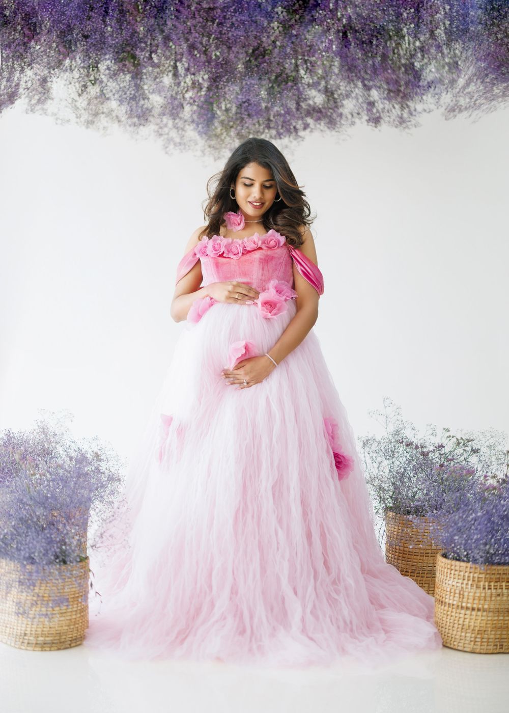 Romantic pink tulle maternity gown with floral details photographed against purple wisteria backdrop.