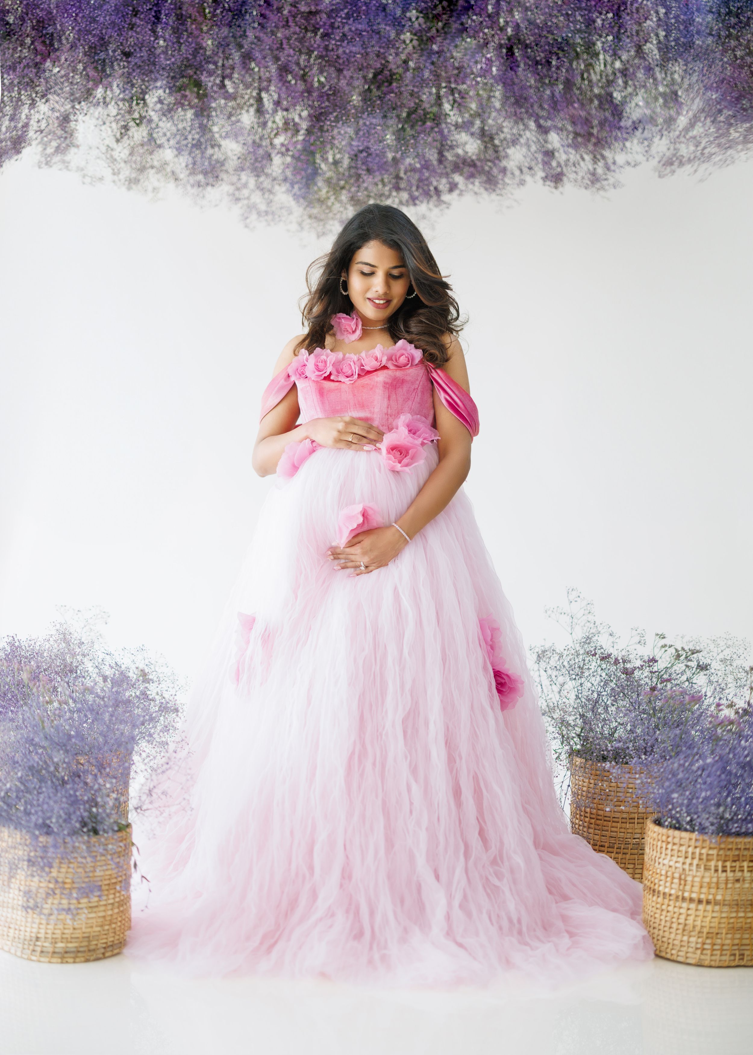 Romantic pink tulle maternity gown with floral details photographed against purple wisteria backdrop.