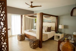 Modern luxury hotel bedroom with four-poster bed, white linens, wooden accents, and decorative woven ottomans.