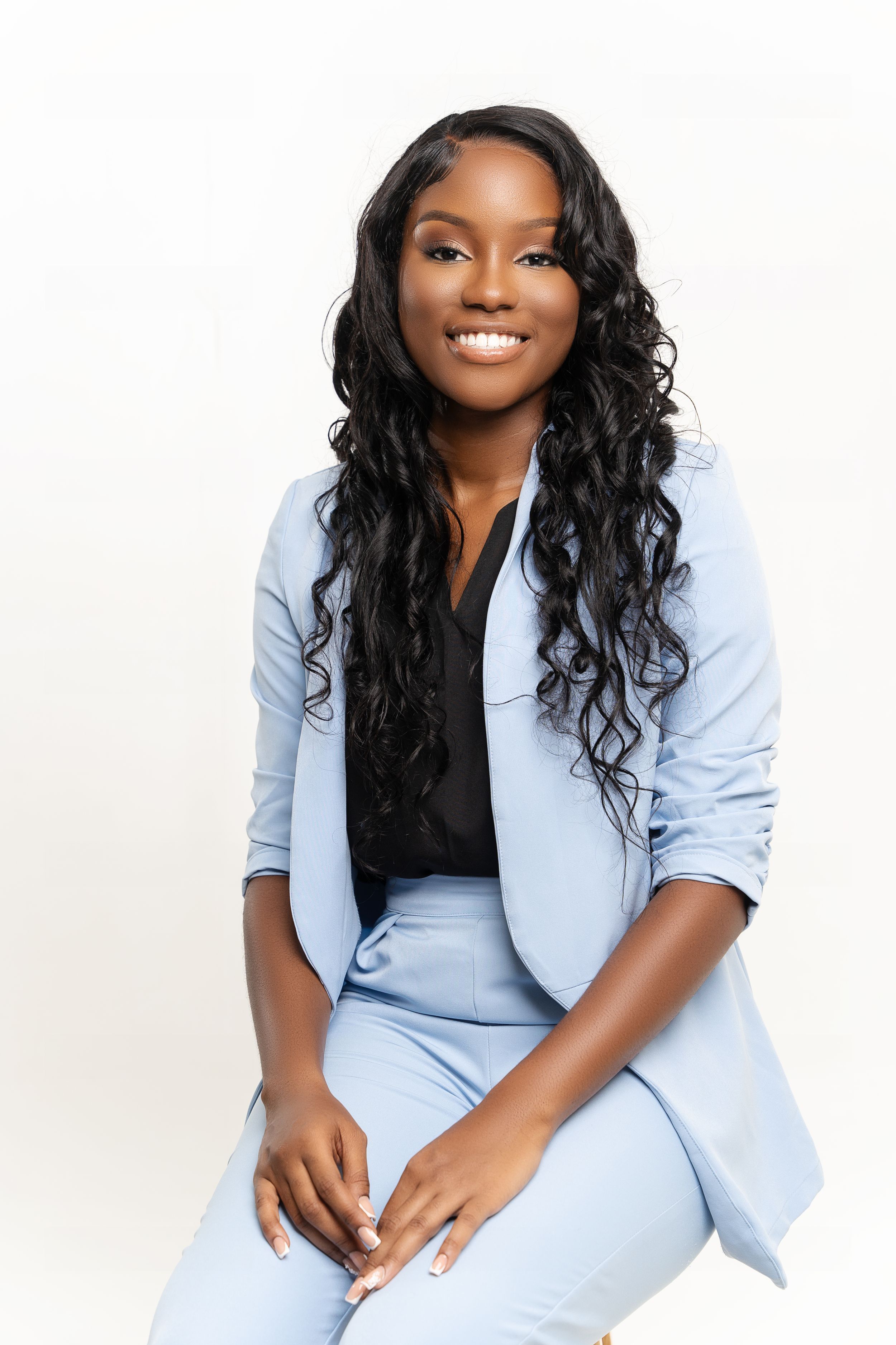 Professional portrait of someone in a light blue pantsuit with long wavy black hair seated against a white background.