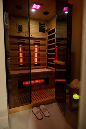 Interior view of a modern infrared sauna cabin with red heating panels, wooden benches, and slippers at the entrance.