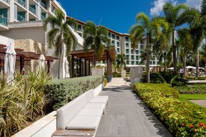 Tropical resort walkway lined with palm trees and landscaping leads to modern hotel entrance under sunny blue skies.