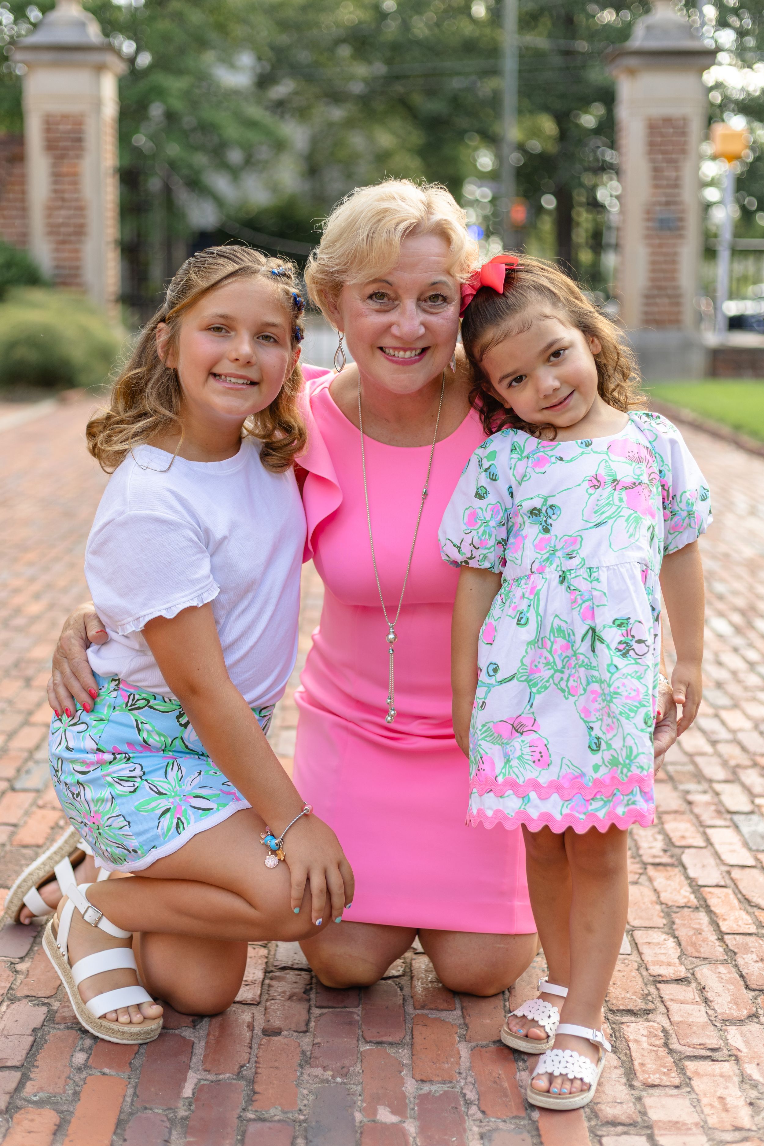 Three people sitting together on brick pavement wearing coordinated pink and floral summer outfits.