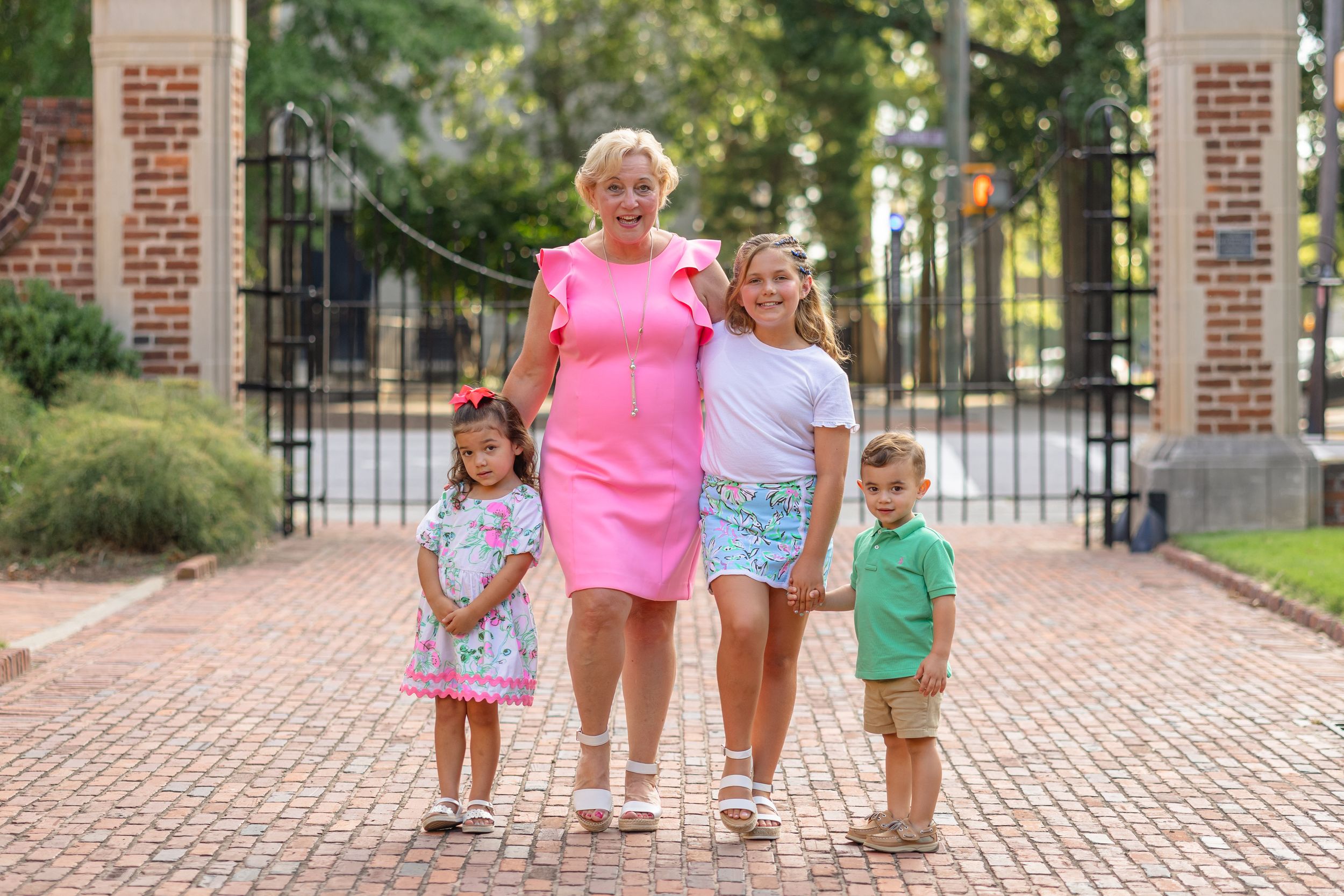 Children walk and play together on a brick pathway near ornate iron gates on a sunny day.