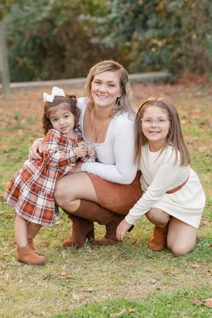 Three children lay on grass outdoors wearing coordinated plaid and white outfits in autumn setting.