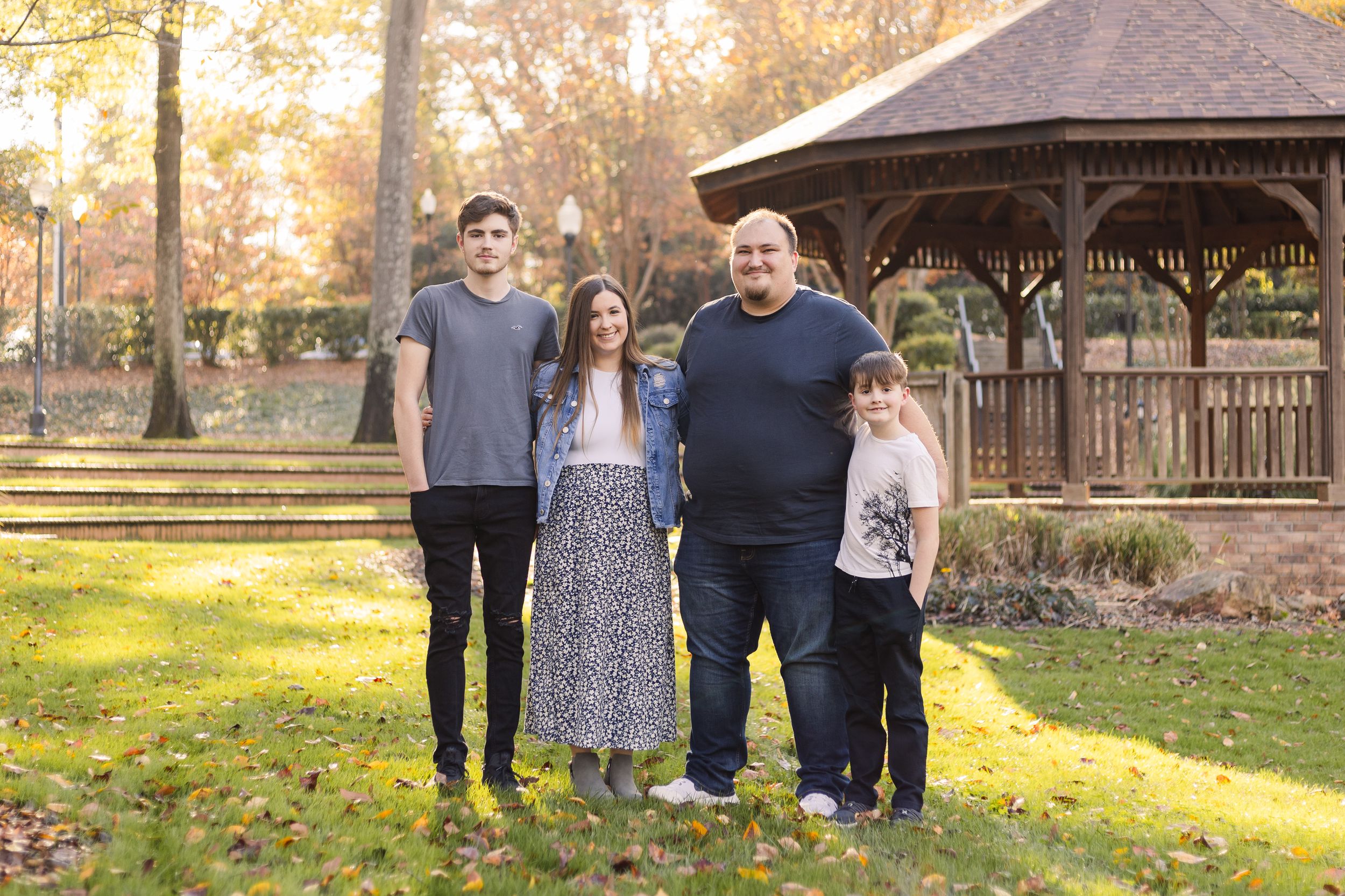 A group stands together on grass in front of a wooden gazebo during autumn with golden sunlight filtering through trees.