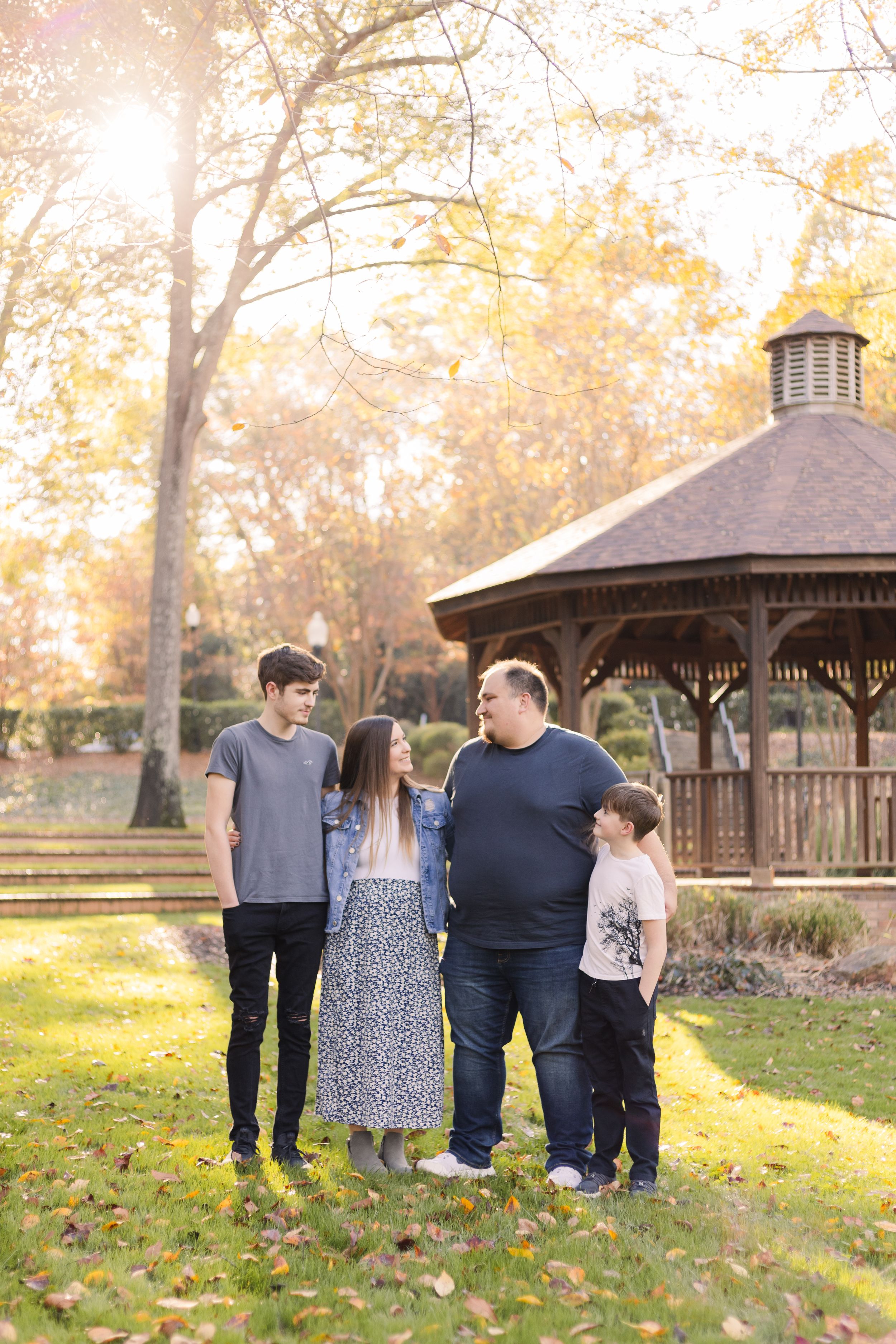 A family poses together in a park during autumn with a rustic gazebo and fallen leaves in the background.