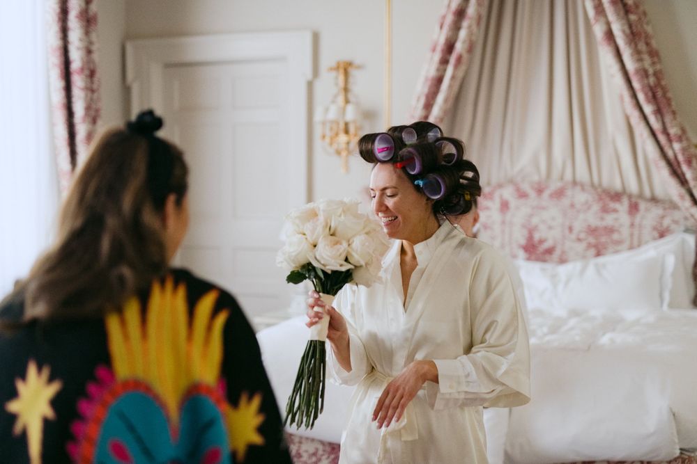 A wedding preparation scene shows someone in a white robe with hair curlers holding a bouquet in an elegant bedroom.
