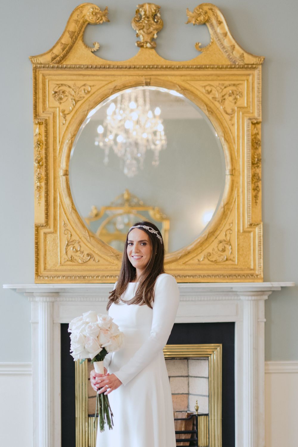 A bride in a white long-sleeved dress poses in front of an ornate gold-framed mirror with a crystal chandelier reflection.