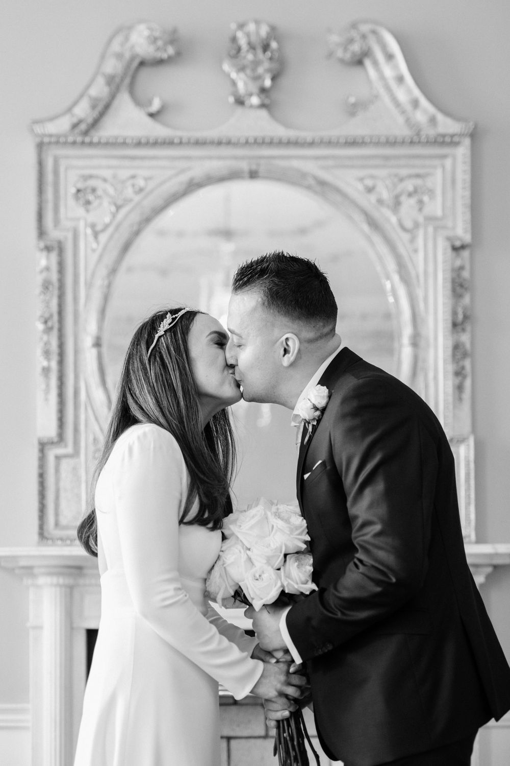 Wedding couple shares a romantic kiss in front of an ornate white marble archway while holding a bouquet of white roses.