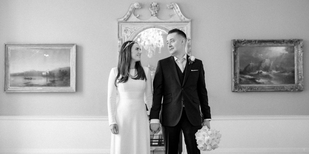 A black and white photograph of a couple holding hands in a formal room with framed paintings on the walls.