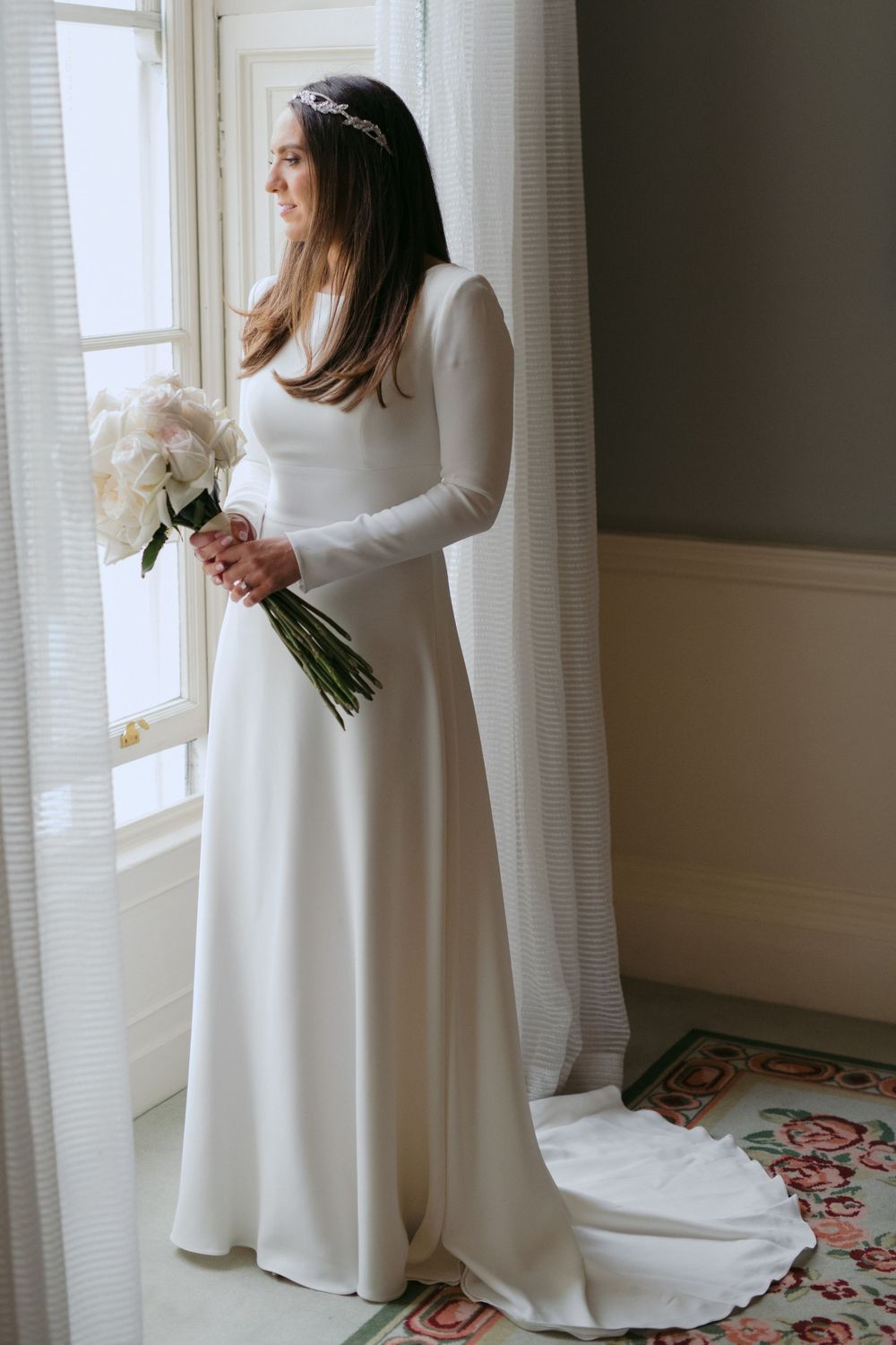 Classic white long-sleeved wedding dress with train photographed by window as figure holds white floral bouquet.