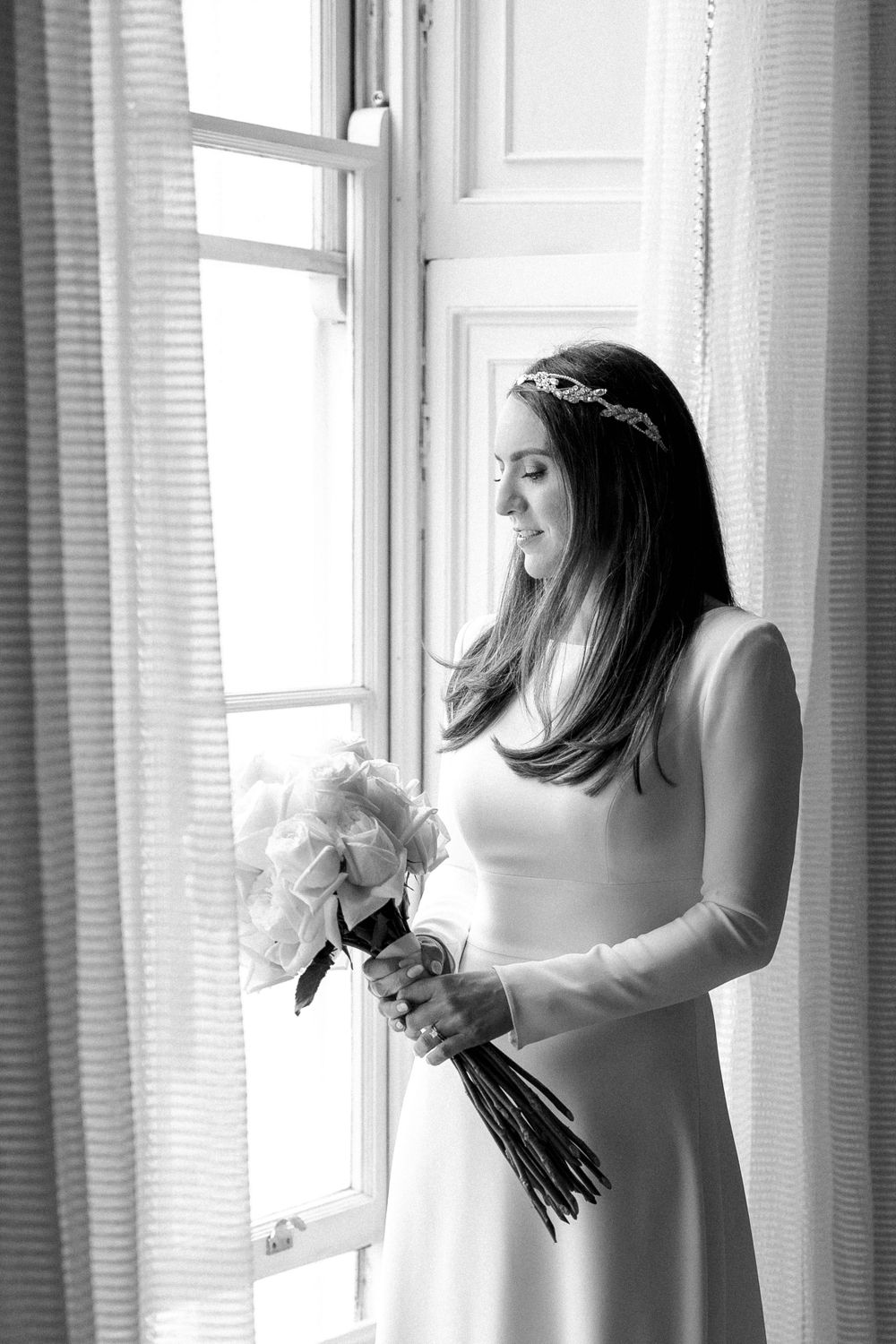 A woman in a white wedding dress stands by a window holding a bouquet of white flowers in black and white photography.