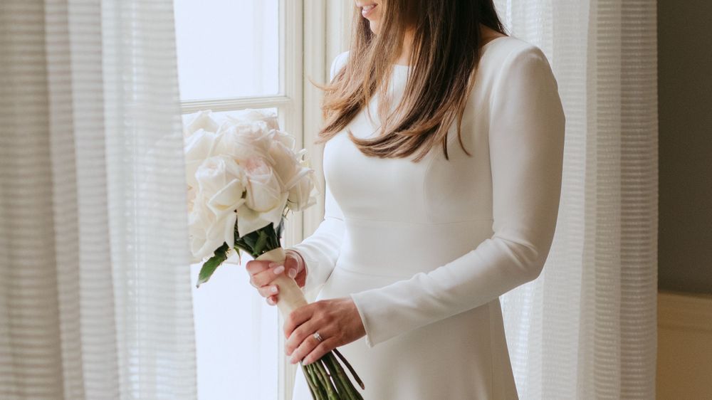 A person in a white long-sleeved dress holds a bouquet of cream-colored peonies while standing by a window.
