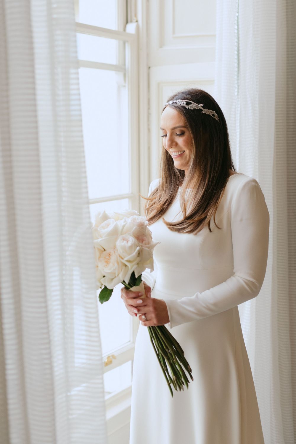 A bride in a classic long-sleeved white wedding dress and delicate headpiece holds a bouquet of cream-colored flowers by a window.