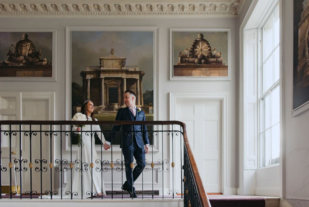 Two figures stand together in an elegant historic interior hallway with ornate moldings and framed artwork on white walls.