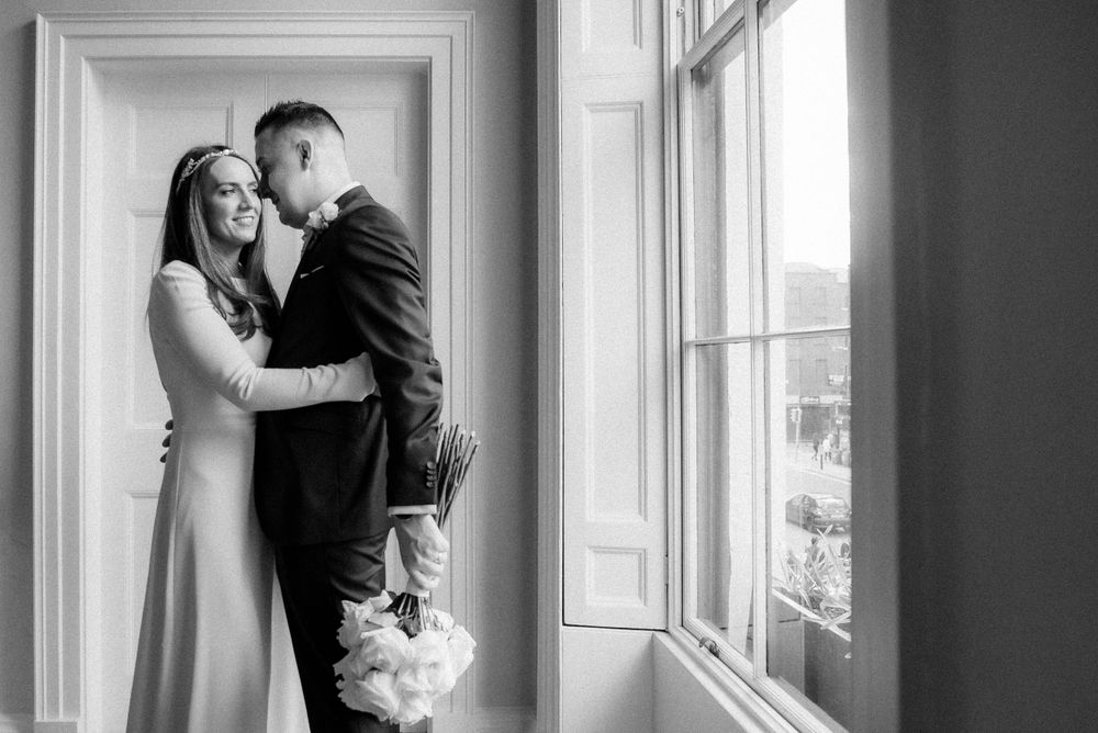 A romantic black and white wedding photograph showing a couple embracing near a bright window.