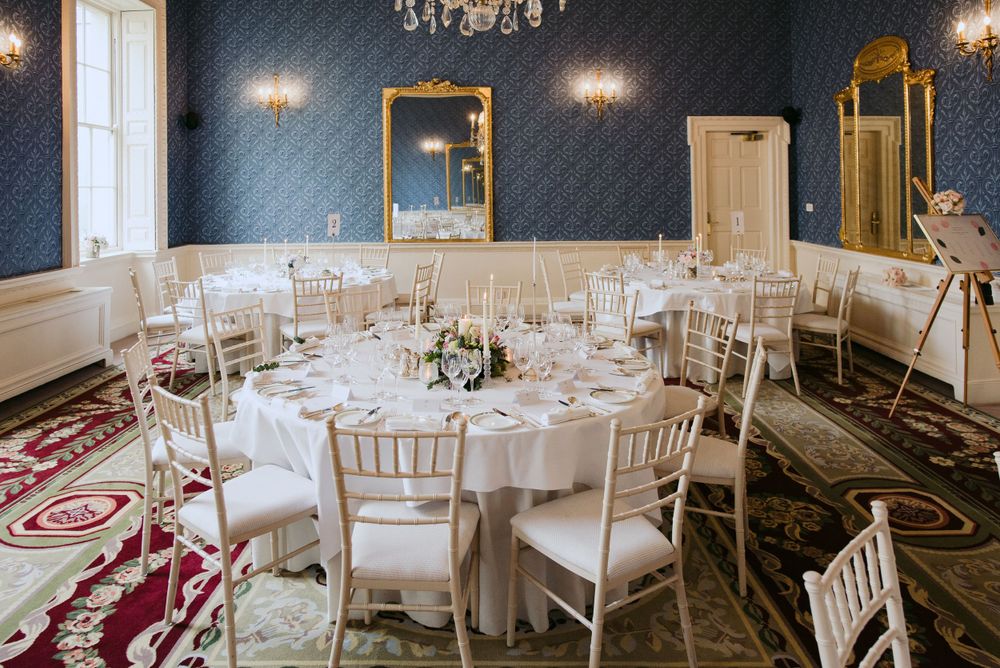 Elegant wedding reception room with navy walls, white chairs and round tables set for dining under crystal chandeliers.