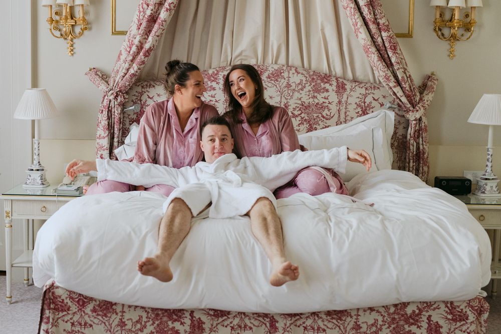 Two people in pink pajamas share laughter while sitting on an elegant canopy bed with toile fabric and white bedding.