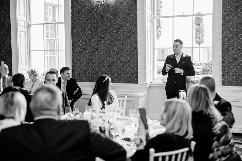 A person delivers a speech during a formal dinner reception in an elegant room with patterned wallpaper and windows.