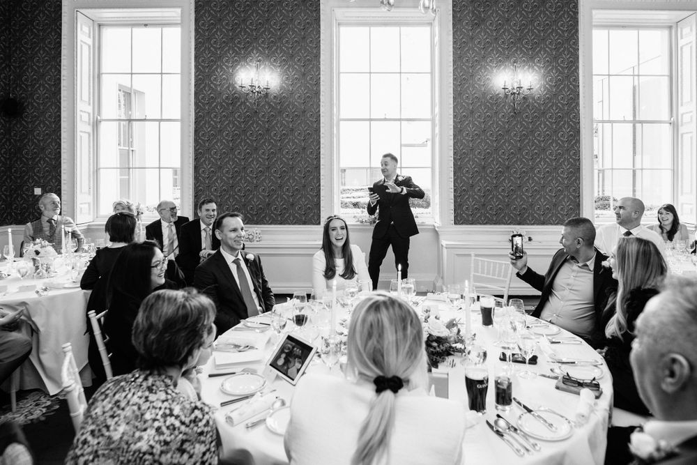 A speaker addresses guests at round tables during an elegant black and white formal dining event in a room with tall windows.
