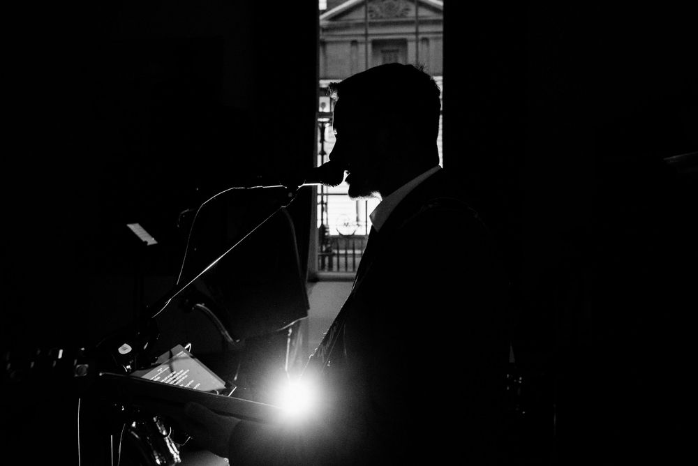 A dramatic silhouette of a person speaking at a microphone against a backlit stage during a performance.