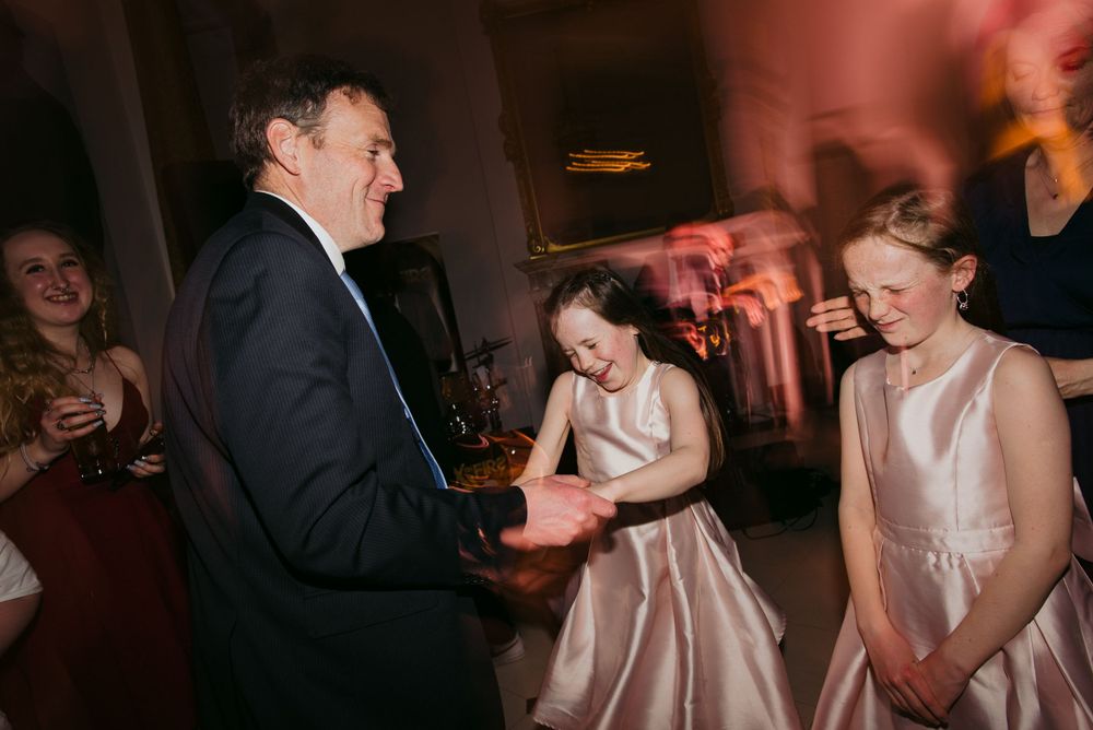 People dancing and having fun at a wedding reception in dimly lit venue with pink lighting.