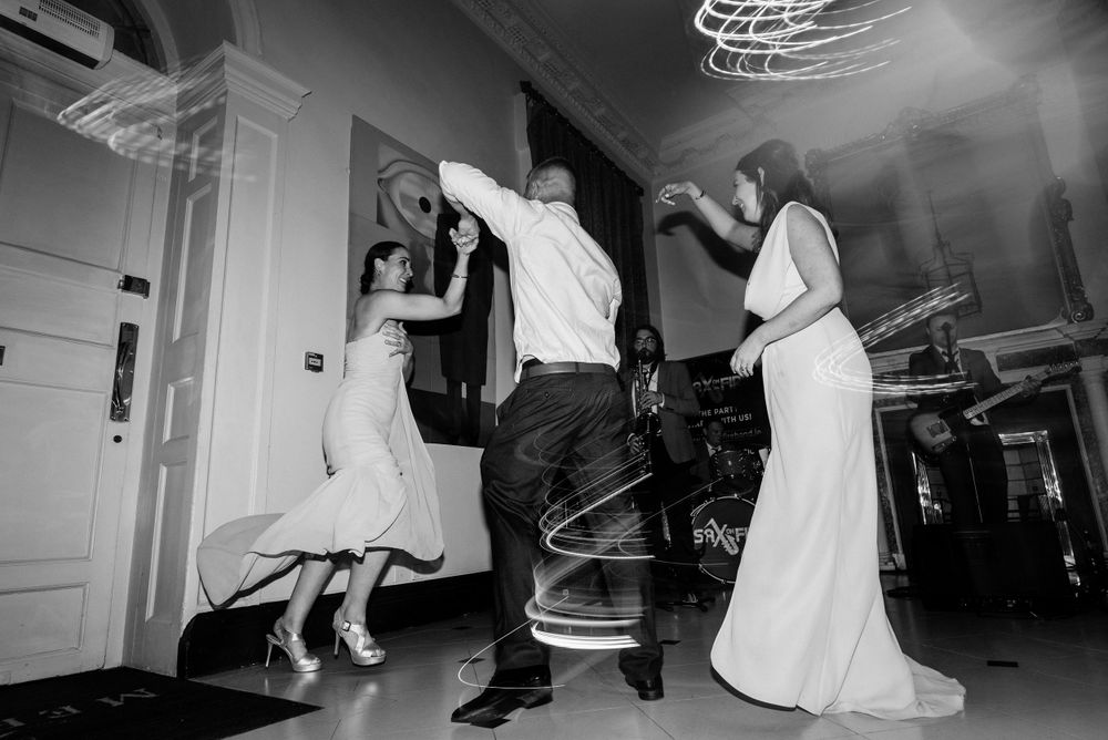 Black and white photo of people dancing and jumping energetically at a wedding reception party indoors.