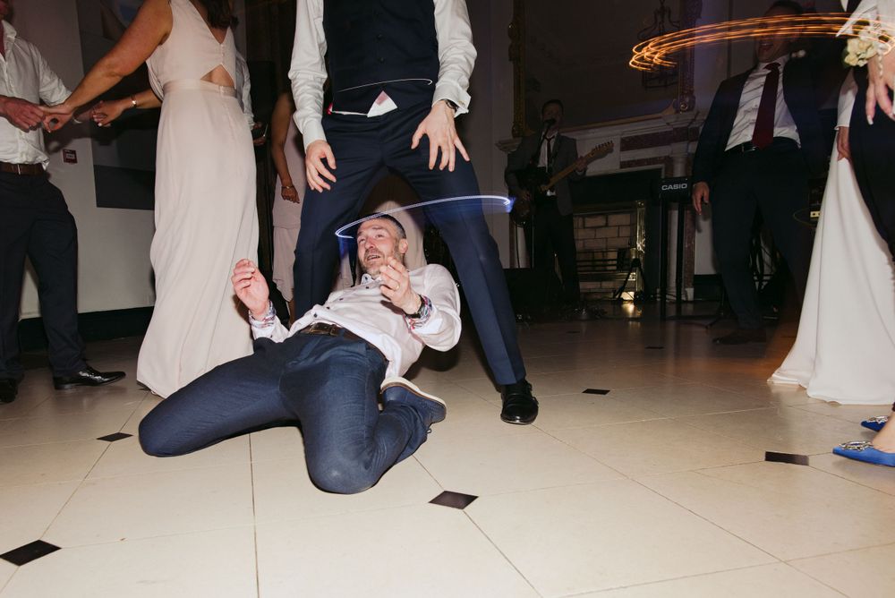 A wedding guest dances enthusiastically on a tiled dance floor, dropping low in jeans and formal attire at a reception.