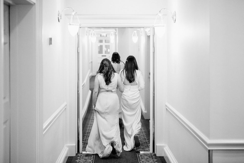 Two people in white wedding dresses walk together down a brightly lit hallway.