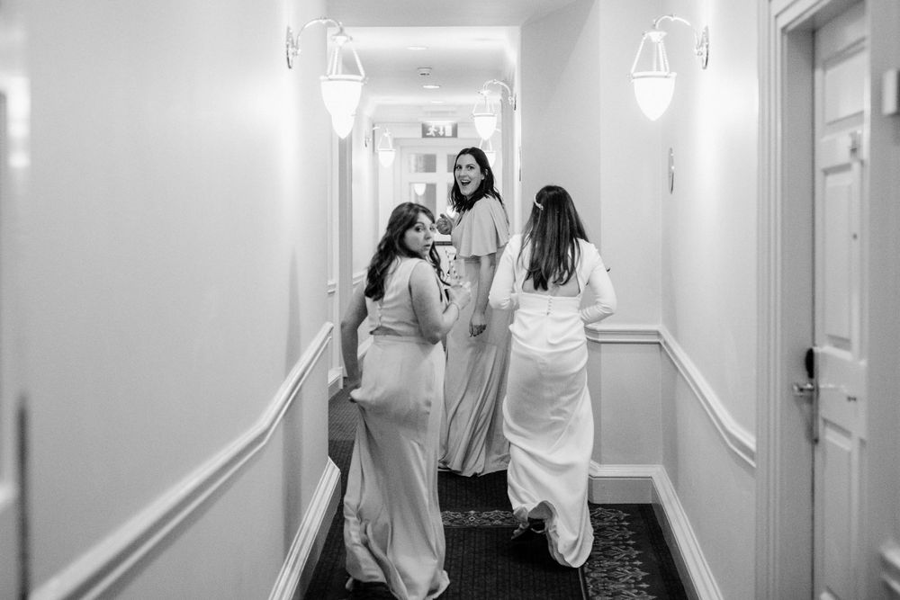 Three people in flowing white dresses walking and interacting in an elegantly lit hotel hallway.
